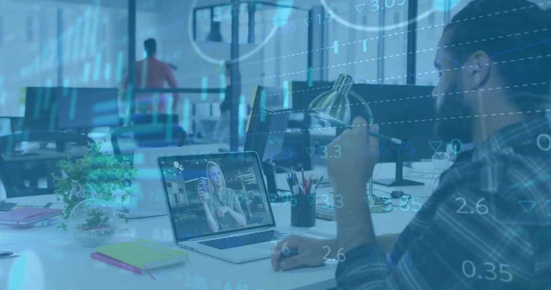 Man holding pen watching remote colleague on laptop with financial data overlay in office