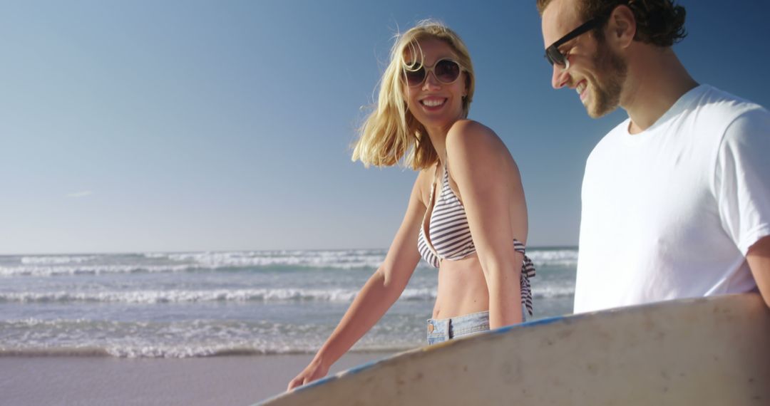 A young Caucasian woman and man carry a surfboard on a sunny beach, with copy space - Free Images, Stock Photos and Pictures on Pikwizard.com