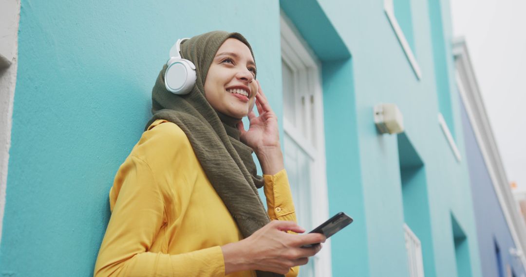 Smiling woman in hijab enjoying music with headphones outside bright turquoise building - Free Images, Stock Photos and Pictures on Pikwizard.com