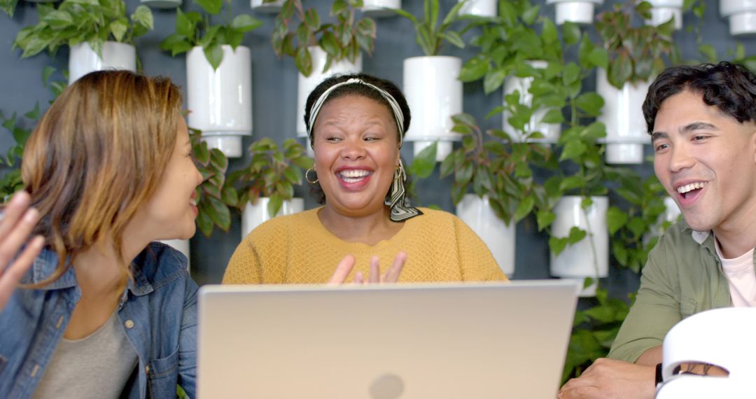Diverse team laughing and collaborating around laptop with plant wall for modern coworking