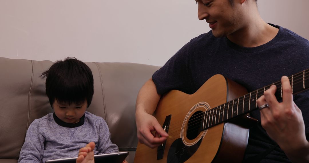 Father Playing Guitar While Son Engaged with Tablet - Free Images, Stock Photos and Pictures on Pikwizard.com