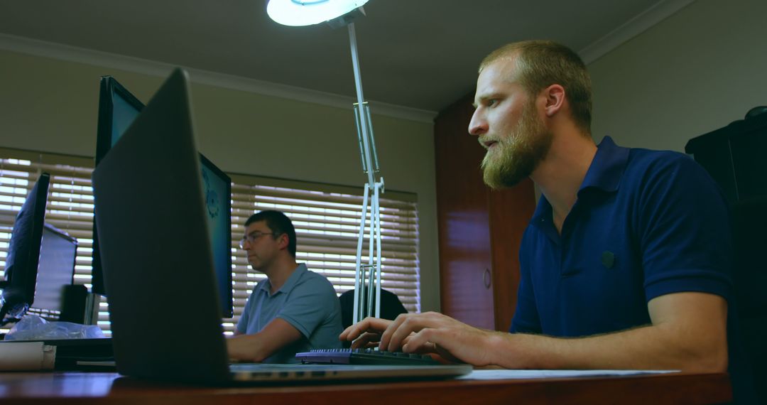 Two Men Working on Laptops in Office Setting with Focus on Bearded Man - Free Images, Stock Photos and Pictures on Pikwizard.com