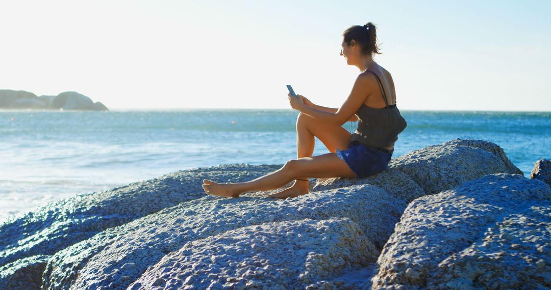 Woman Relaxing on Coastal Rocks Using Smartphone During Summer Day - Free Images, Stock Photos and Pictures on Pikwizard.com