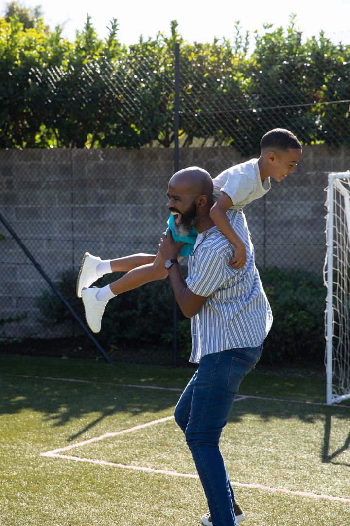 Father Carrying Child Piggyback on Backyard Soccer Pitch Joyfully