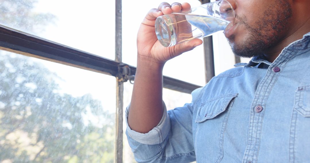 Man in Denim Drinking Water by Window for Refreshment and Hydration