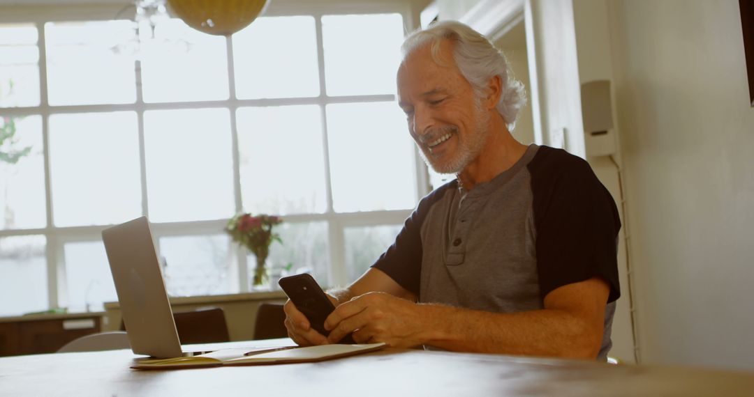 Senior Man Using Smartphone While Working on Laptop in Bright Home Office - Free Images, Stock Photos and Pictures on Pikwizard.com