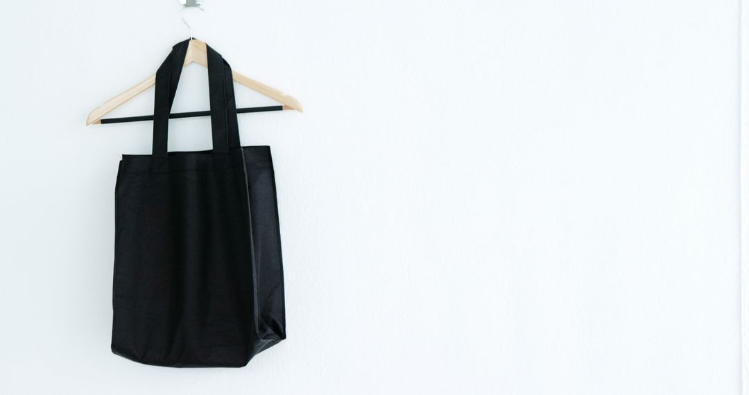 A black tote bag hangs on a wooden hanger against a white wall, with copy space - Free Images, Stock Photos and Pictures on Pikwizard.com
