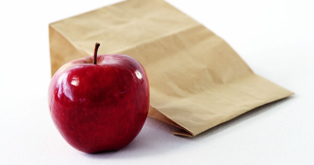 A shiny red apple sits in front of a brown paper bag on a white surface, with copy space - Free Images, Stock Photos and Pictures on Pikwizard.com