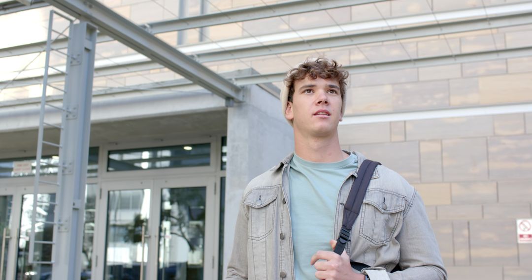 Young commuter standing outside modern campus building carrying backpack and looking ahead