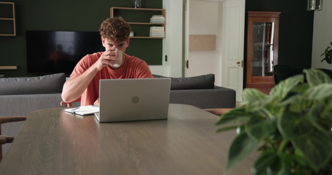 Young man working from home at wooden table sipping coffee laptop remote workspace modern