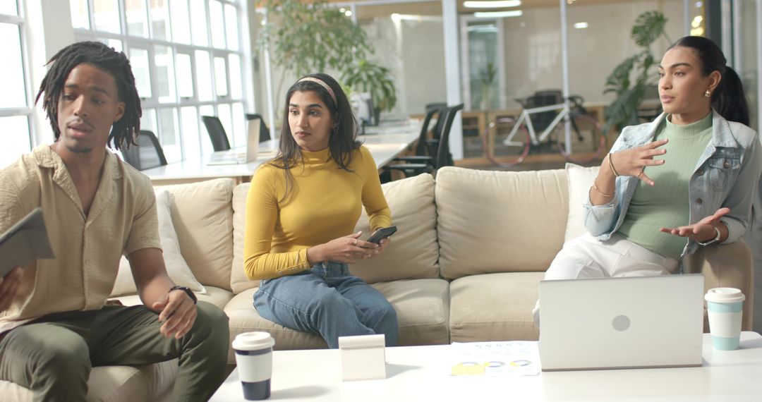 Diverse coworkers discussing project on lounge sofa with laptop, coffee, casual teamwork