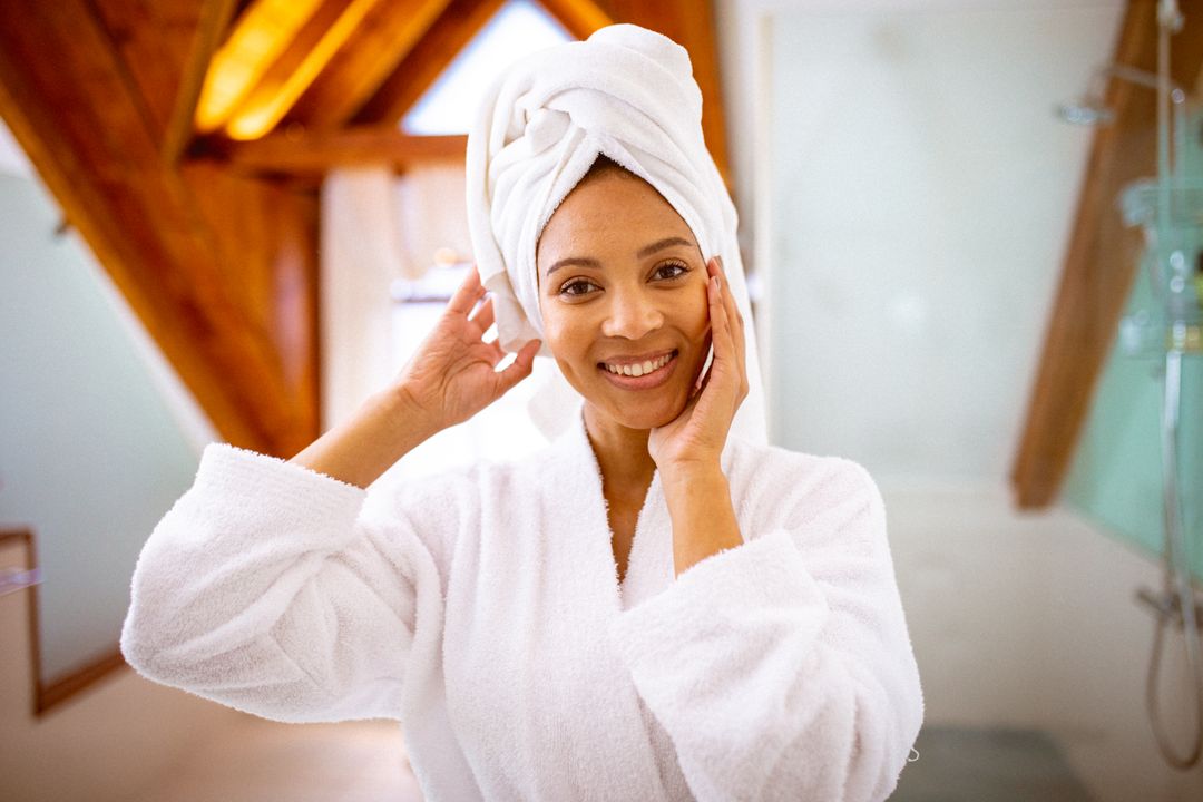 Smiling biracial woman wearing robe and towel turban looking in mirror - Free Images, Stock Photos and Pictures on Pikwizard.com