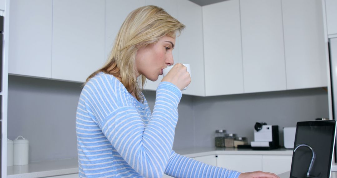 Blonde Woman Drinking Coffee and Working on Laptop in Modern Kitchen - Free Images, Stock Photos and Pictures on Pikwizard.com