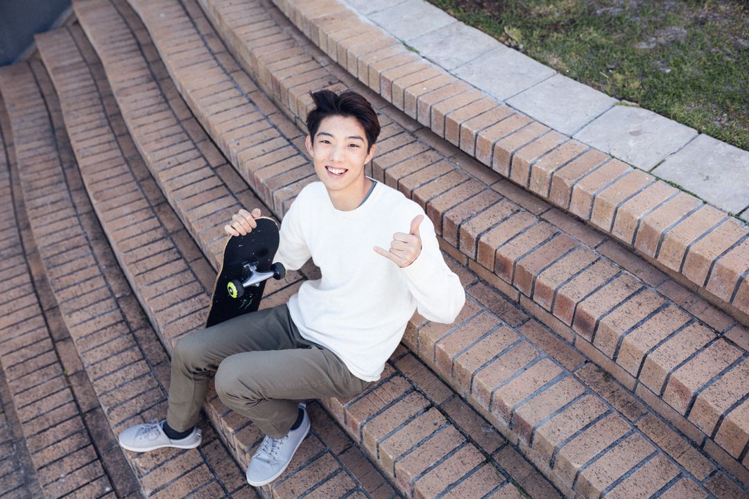 High angle portrait of smiling asian teenage boy gesturing while sitting with skateboard on steps - Free Images, Stock Photos and Pictures on Pikwizard.com