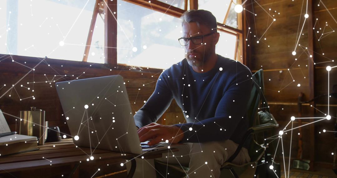 Mature man working remotely in rustic cabin typing on laptop with digital network overlay
