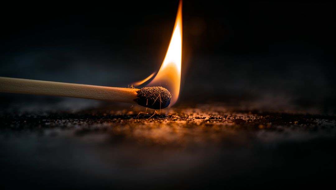 Burning matchstick head glowing with flame and embers on dark textured surface