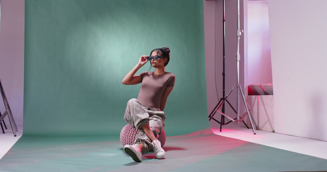 Female model adjusting sunglasses while sitting on pink pouf against green studio backdrop