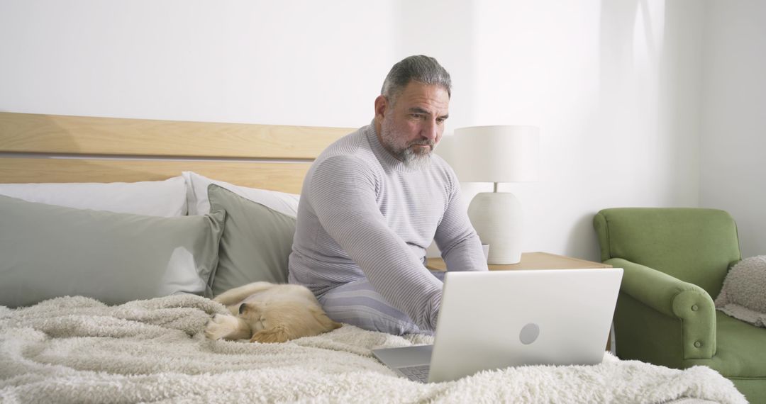 Bearded man in grey loungewear working on laptop with sleeping dog