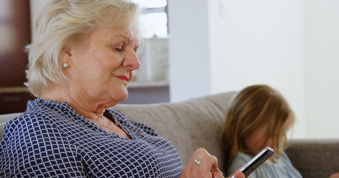 Elderly woman using smartphone with granddaughter playing in background - Free Images, Stock Photos and Pictures on Pikwizard.com