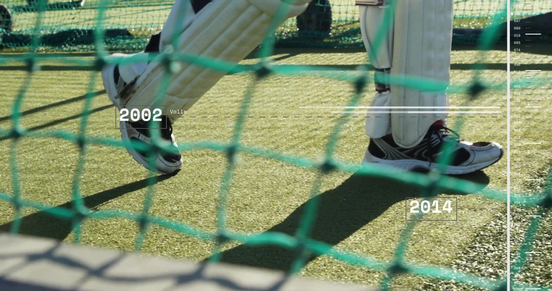 Framing batter wearing white batting pads and sneakers through green netting on turf