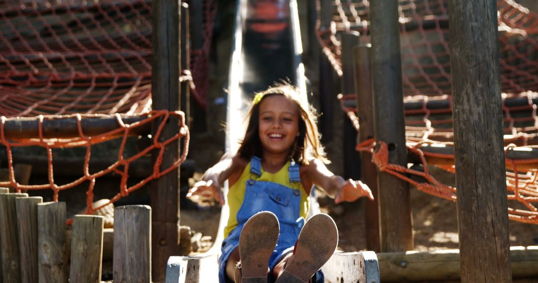 Smiling Girl in Blue Overalls Sliding Down Playground Slide on Sunny Day - Free Images, Stock Photos and Pictures on Pikwizard.com