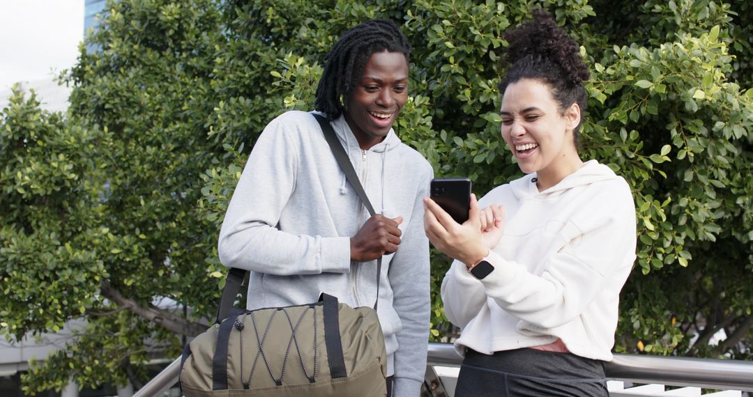 Diverse students laughing while checking smartphone, carrying duffel by campus railing