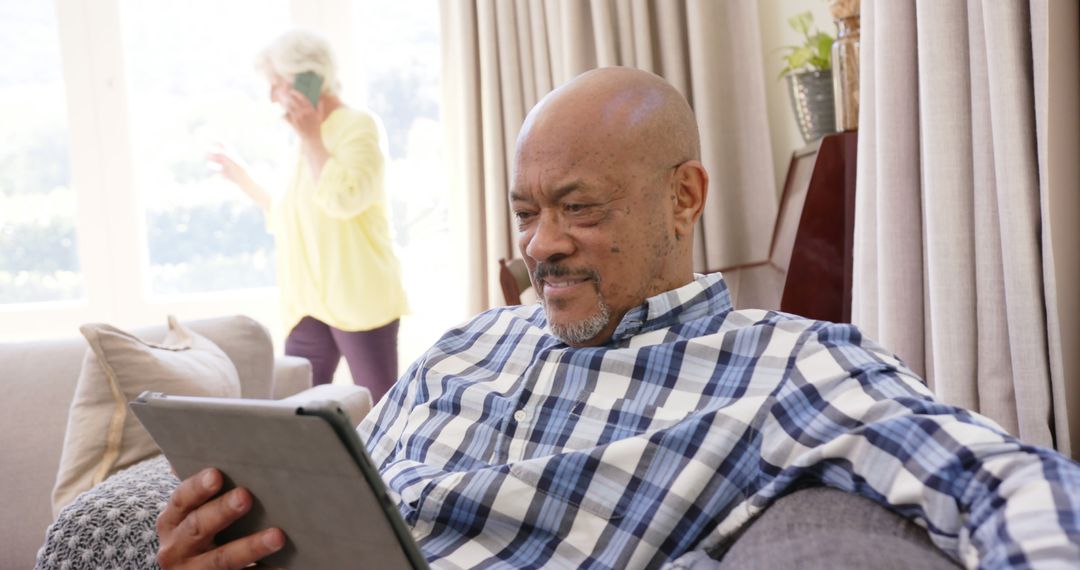 Senior Man Using Tablet at Home While Woman in Background is on Phone Call - Free Images, Stock Photos and Pictures on Pikwizard.com