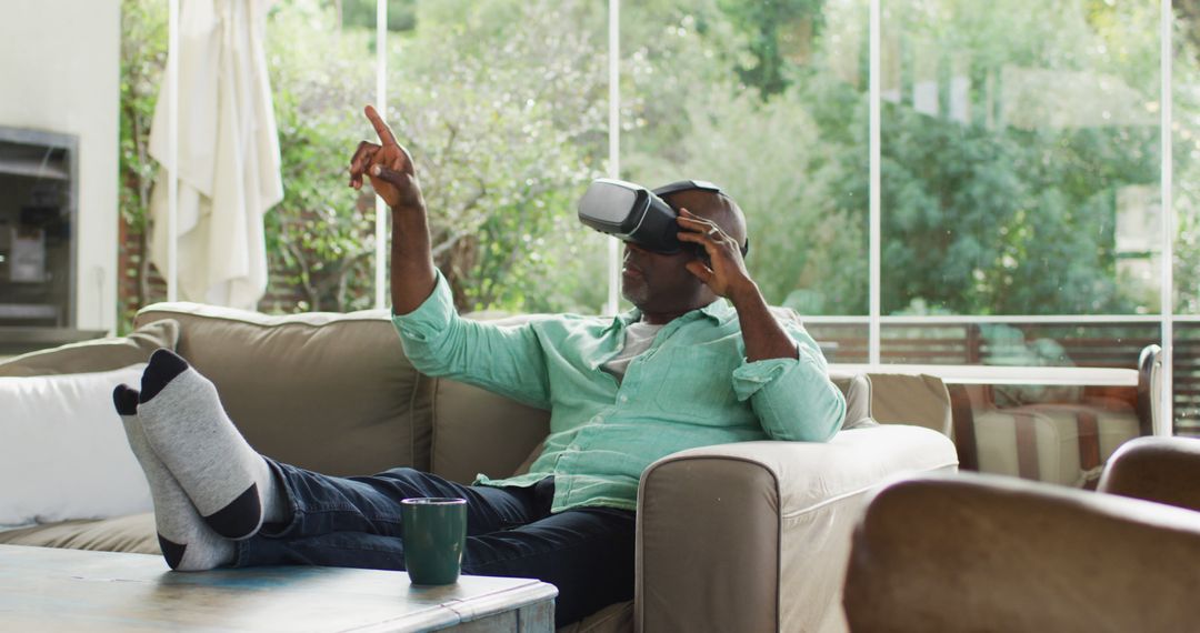 Happy african american senior man sitting on couch with feet up using vr headset and pointing - Free Images, Stock Photos and Pictures on Pikwizard.com