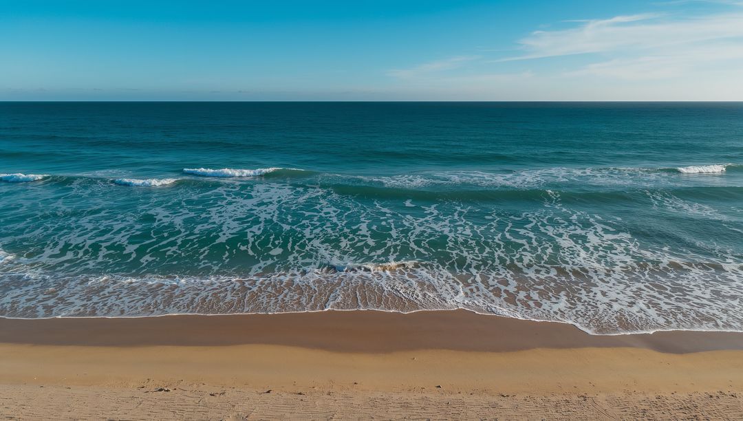 Aerial view of turquoise waves lapping golden sandy beach with foamy surf and clear horizon