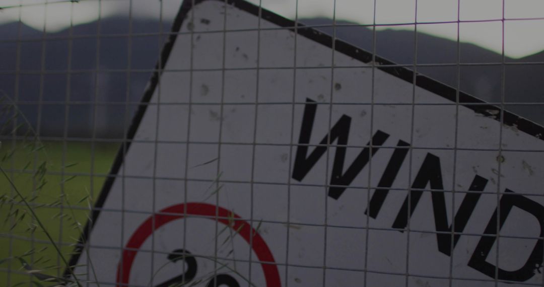 Leaning weathered wind warning sign behind wire mesh with tall grass in rural field