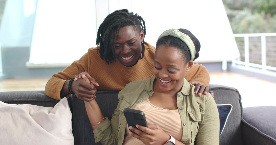 African American couple relaxing on sofa sharing joyful moment while using smartphone