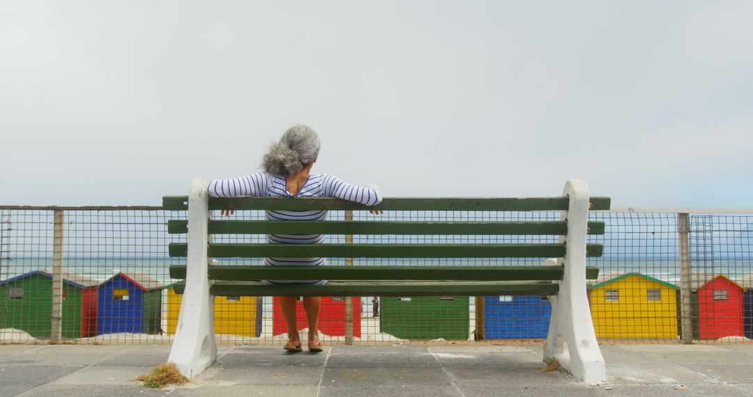 Senior Woman Relaxing on Bench Overlooking Colorful Beach Huts