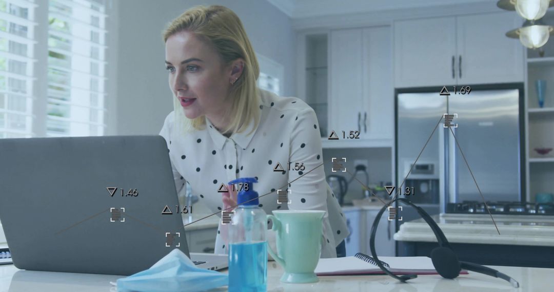 Woman working from home in modern kitchen leaning over laptop with headset and face mask
