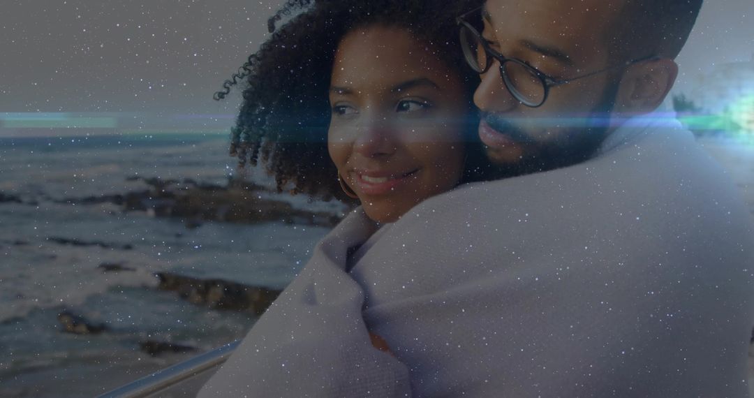 Embracing couple snuggling on seaside railing wrapped in blanket at dusk with lens flare