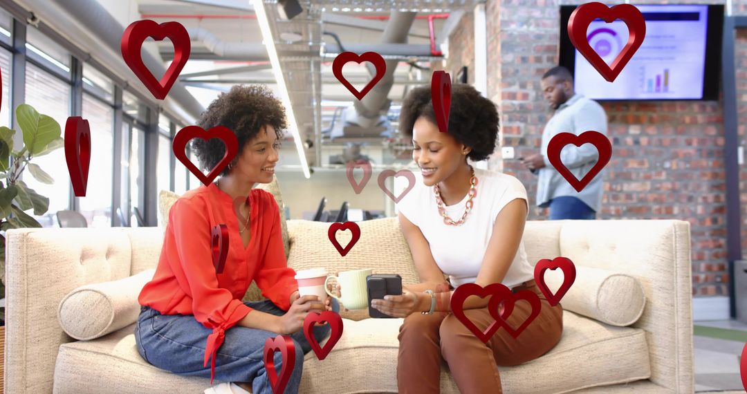 Two coworkers chatting on office sofa holding coffee and device surrounded by red hearts