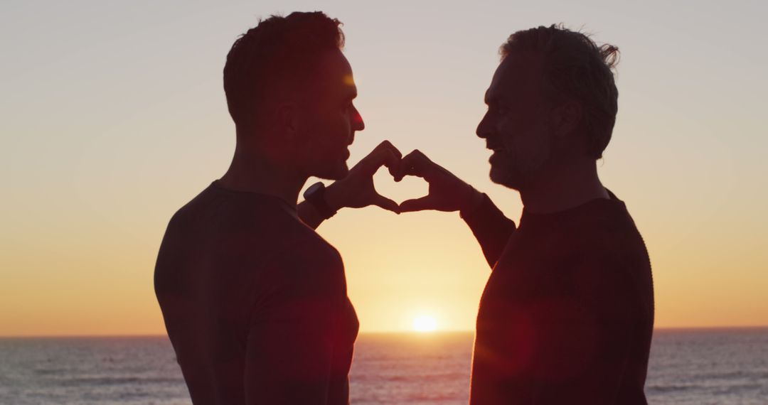 Happy caucasian gay male couple holding hands and making heart shape at sunset on the beach - Free Images, Stock Photos and Pictures on Pikwizard.com