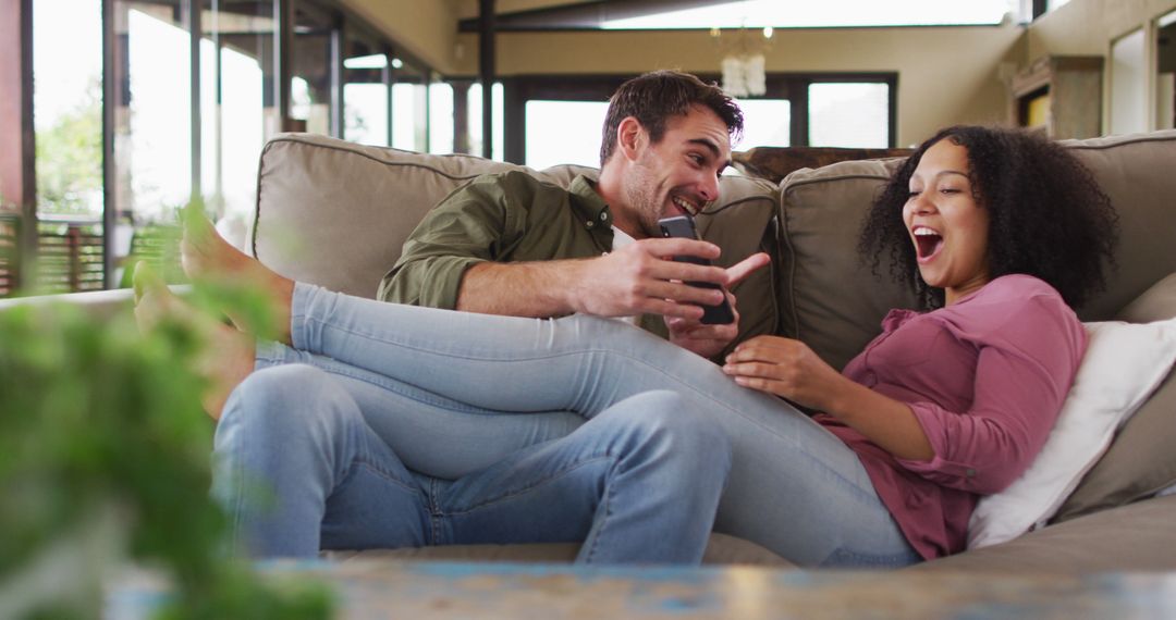 Biracial couple smiling while looking in smartphone together on the couch at vacation home - Free Images, Stock Photos and Pictures on Pikwizard.com