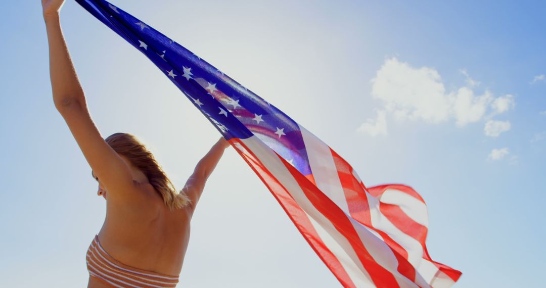 A young Caucasian woman holds the American flag high against a clear sky, with copy space - Free Images, Stock Photos and Pictures on Pikwizard.com