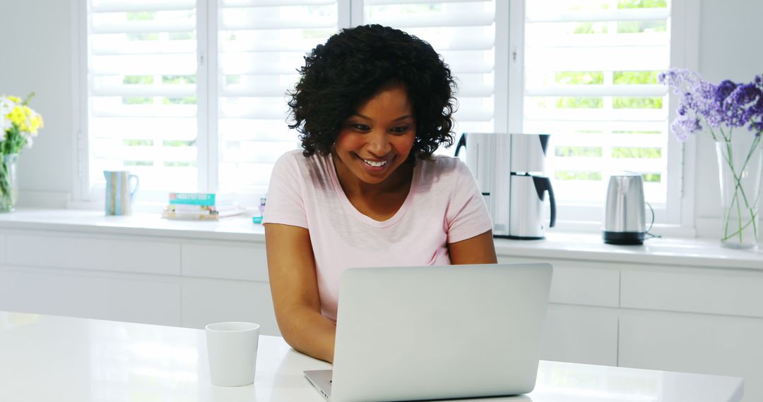 Woman smiling while working on laptop in bright kitchen - Free Images, Stock Photos and Pictures on Pikwizard.com