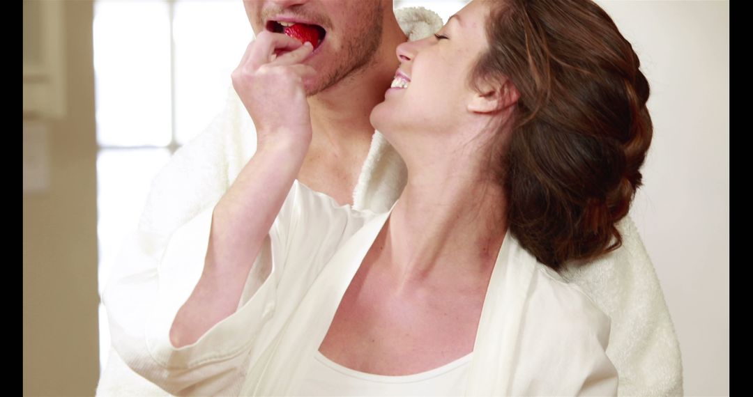 A joyful couple in white bathrobes shares a strawberry, hinting at romance in a cozy setting. - Free Images, Stock Photos and Pictures on Pikwizard.com