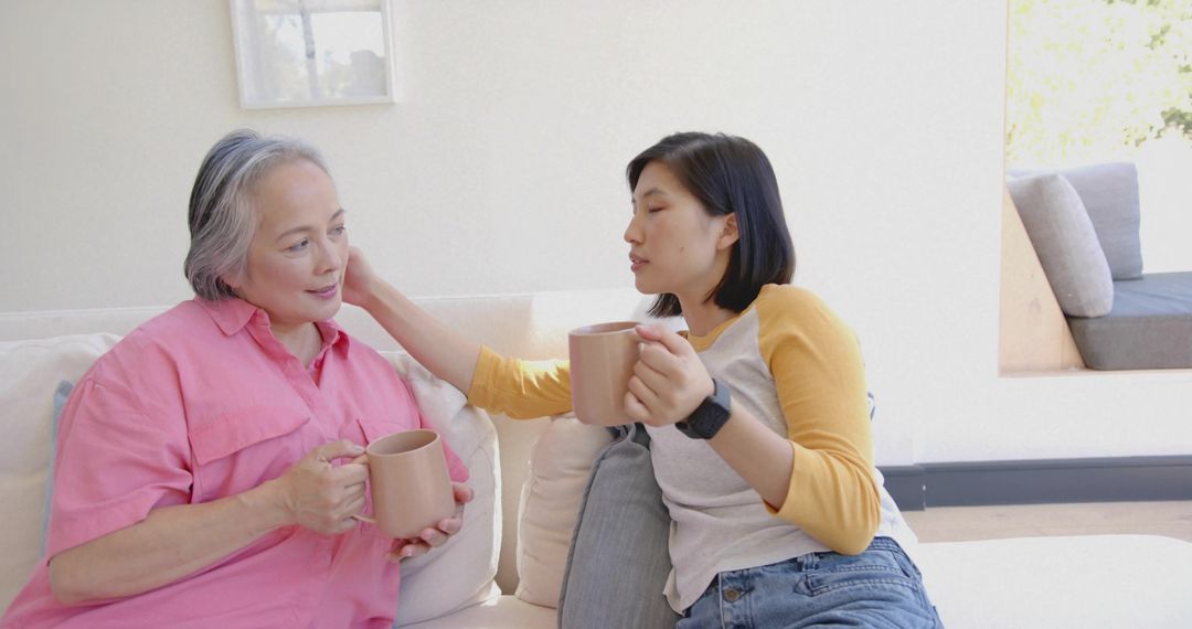 Comforting adult touching senior on sofa sharing warm mugs and gentle conversation at home