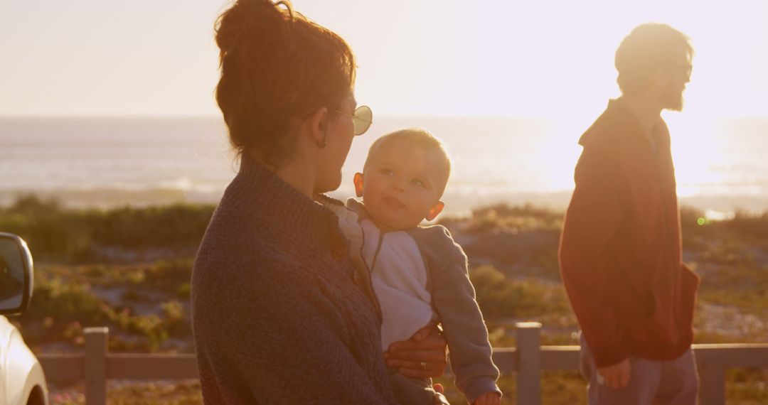 Mother Holding Baby Outdoors at Sunset with Man in Background - Free Images, Stock Photos and Pictures on Pikwizard.com