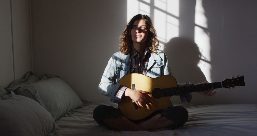 Young Woman Sitting on Bed Playing Acoustic Guitar in Sunlit Bedroom - Free Images, Stock Photos and Pictures on Pikwizard.com