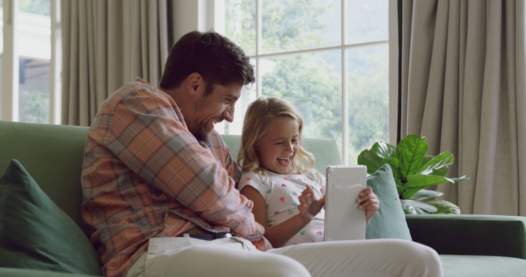 Father and Daughter Laughing Together Using Digital Tablet on Couch - Free Images, Stock Photos and Pictures on Pikwizard.com