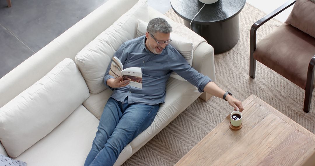 Man Relaxing on Sofa Reading and Reaching for Coffee Mug - Free Images, Stock Photos and Pictures on Pikwizard.com