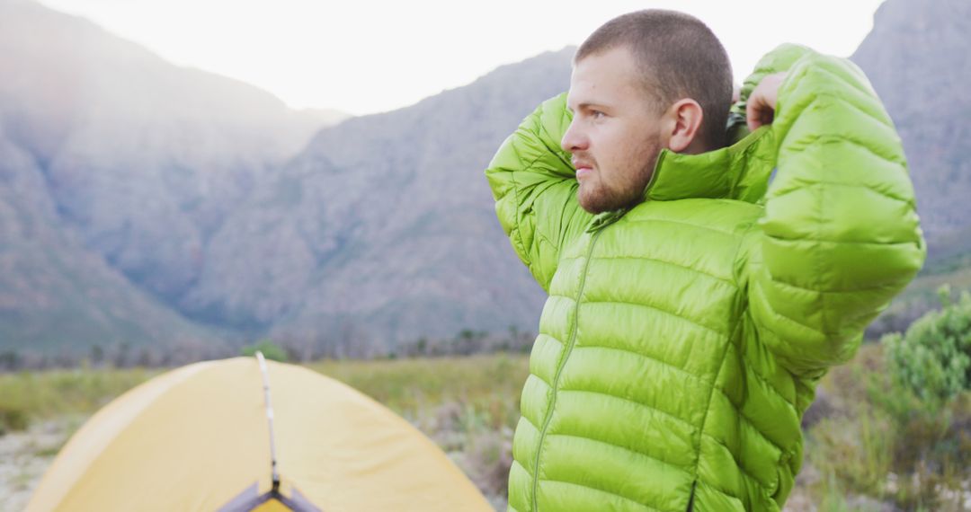 Man Adjusting Green Jacket in Mountain Campsite - Free Images, Stock Photos and Pictures on Pikwizard.com