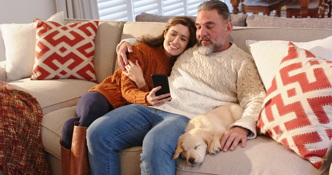 Mature couple cuddling on couch with sleeping puppy while scrolling smartphone
