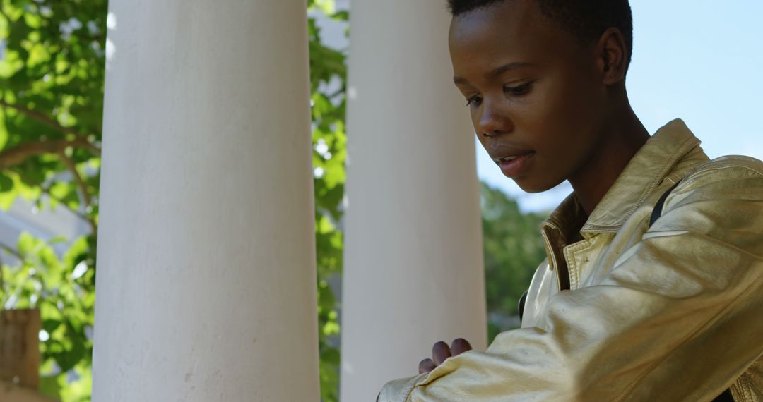 Young person wearing gold jacket standing near white columns and greenery - Free Images, Stock Photos and Pictures on Pikwizard.com