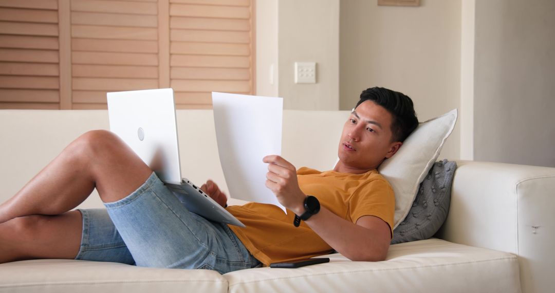 Man Relaxing on Sofa Reviewing Documents with Laptop and Smartphone
