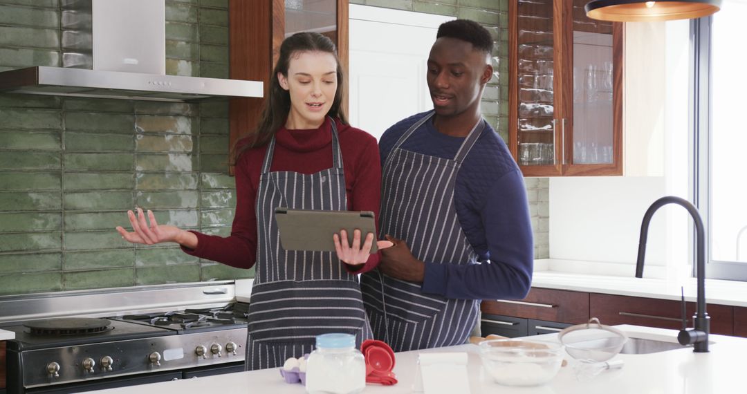 Couple Wearing Aprons Following Recipe on Digital Tablet in Modern Kitchen - Free Images, Stock Photos and Pictures on Pikwizard.com